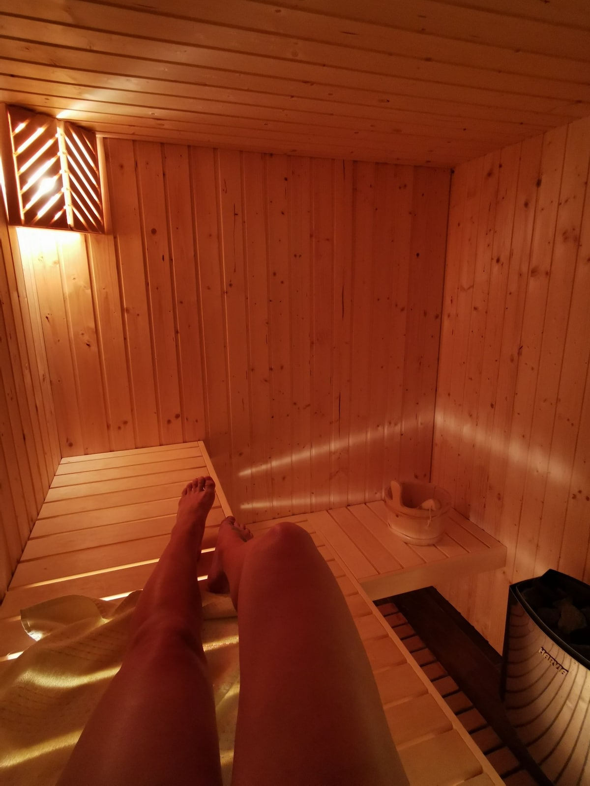 The sauna area features warm wooden paneling and soft lighting, creating a relaxing environment. A wooden bench runs along one side, while a small bucket and ladle are positioned nearby. A waste bin can be seen in the corner, completing the tranquil space.
