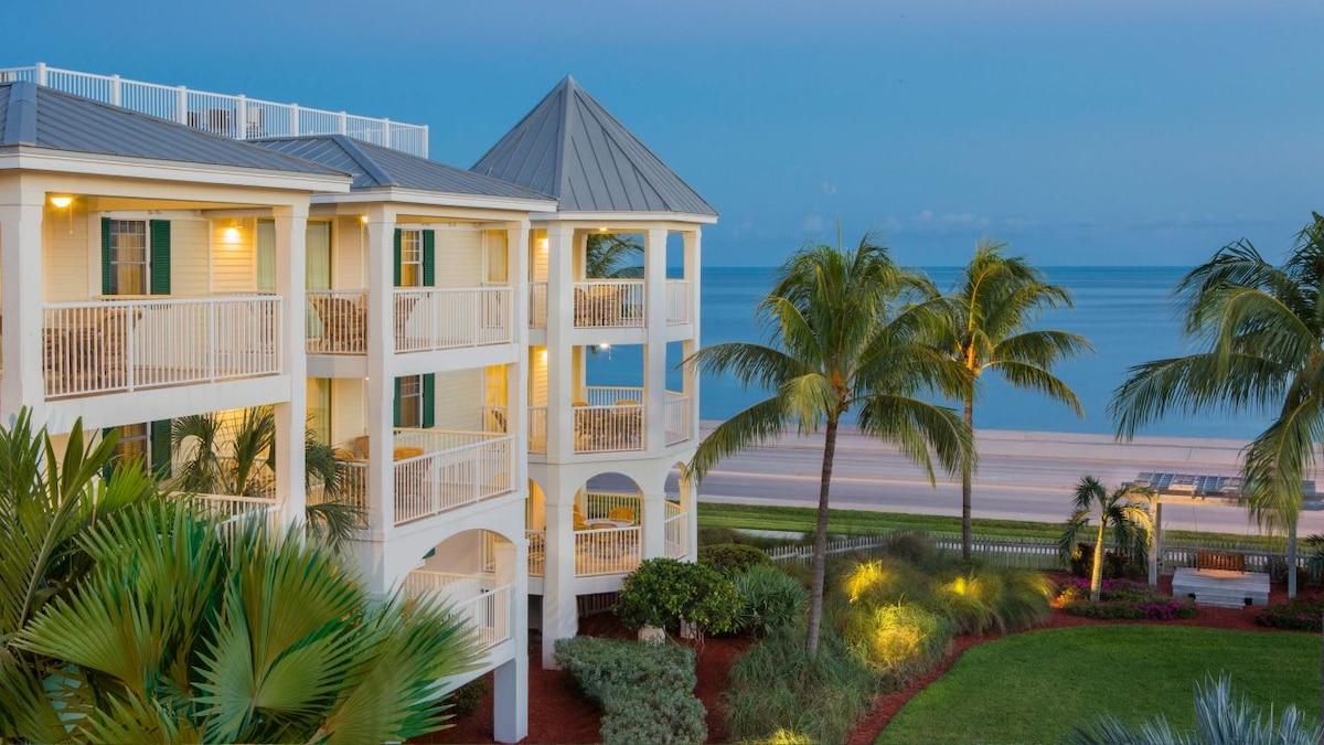 A three-story building with a metal roof stands by the shoreline, featuring multiple balconies lined with white railings. Palm trees are observed around the landscaped grounds, illuminated softly by garden lights, with the ocean visible beyond the beach in the background.