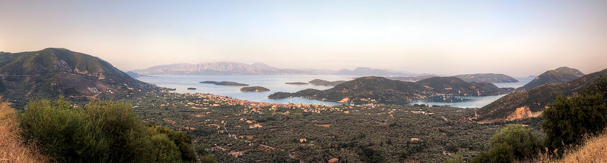 A panoramic view reveals the tranquil landscape of the east side of the island, showcasing the sea dotted with several smaller islands and lush green hills. The soft colors of the sky transition from warm to cool tones, indicating either dawn or dusk.