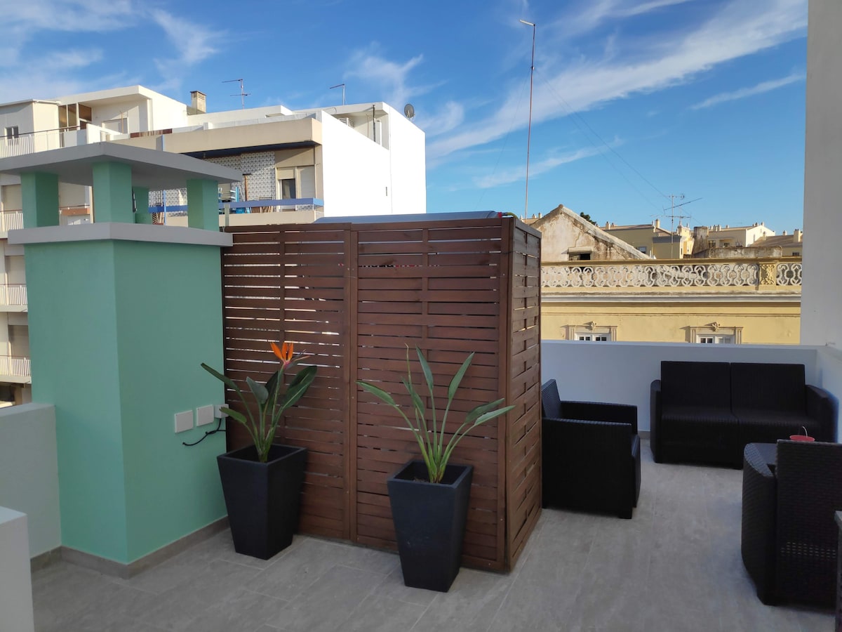 An outdoor terrace features a seating area with two dark wicker chairs and a small table. Lush green plants are positioned in black planters, while a wooden privacy screen adds a partition. A clear blue sky is visible above, contributing to a relaxing atmosphere.