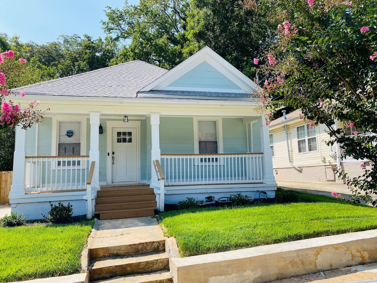 A charming single-story house features a light blue exterior with white trim. A covered porch with white railings invites guests. Lush green grass is seen around the property, complemented by flowering plants, while a stone pathway leads to the front door.