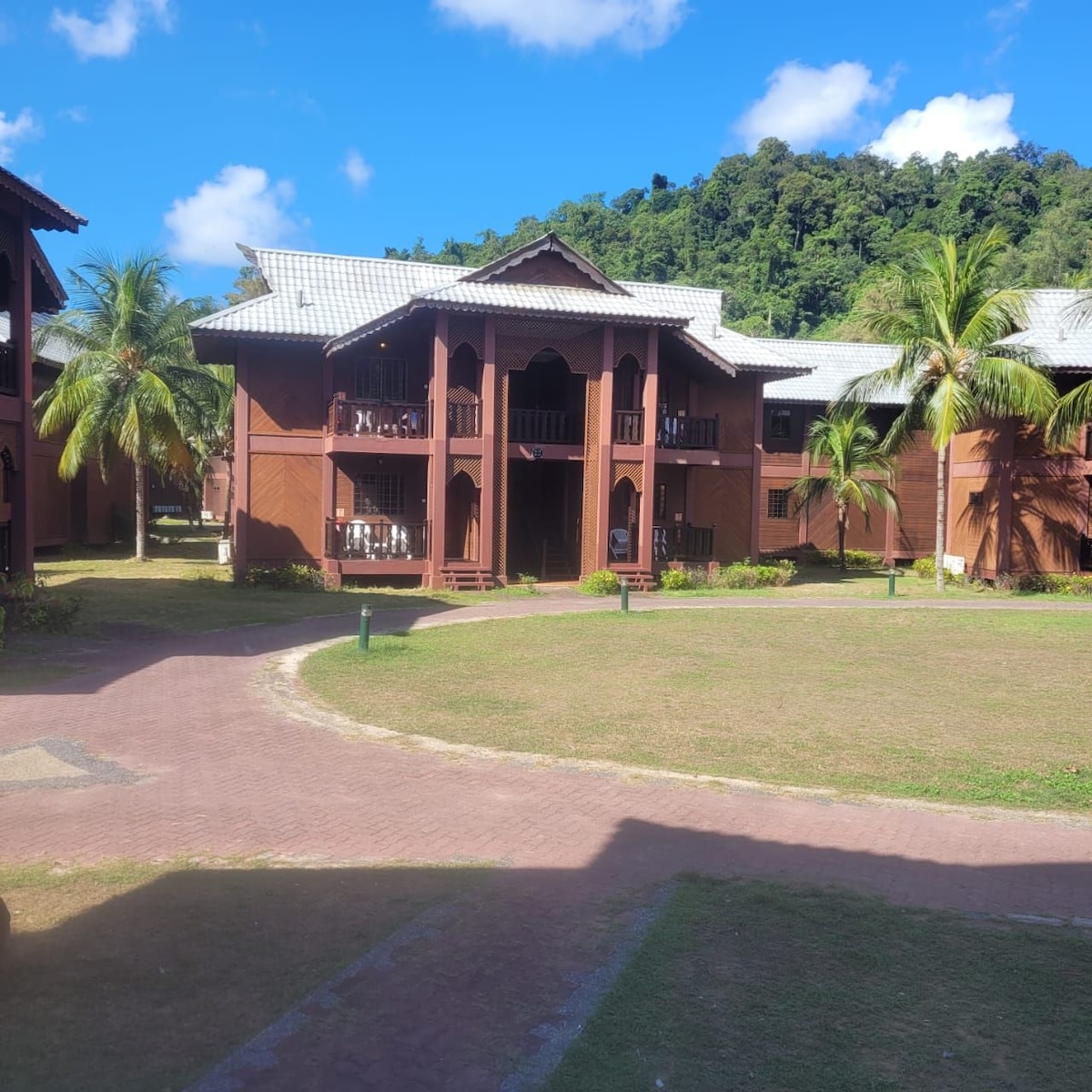 A charming two-story wooden building features a welcoming entrance with a spacious porch. Lush green palm trees surround the exterior, and a well-manicured lawn leads to a walkway. A mountainous backdrop adds to the tranquil setting.