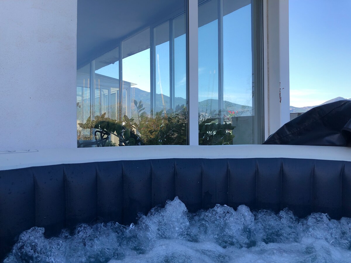 A hot tub with bubbling water is viewed from an outdoor area. The glass walls of the building reflect the surrounding landscape, including mountains and greenery. Natural light illuminates the scene, enhancing the tranquil atmosphere.