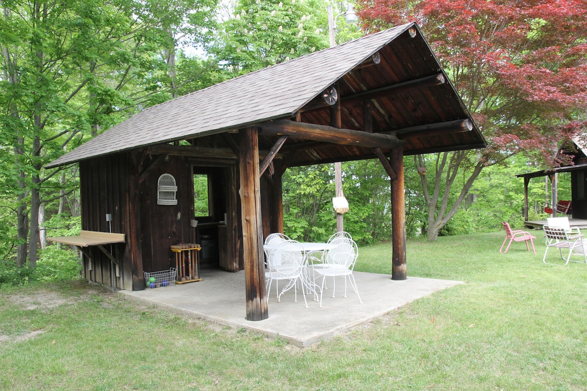 An open-air summer kitchen features a large overhanging roof supported by wooden beams. A concrete patio is furnished with a round table and four white chairs. Nearby, a small wooden countertop and a mailbox are visible, all surrounded by lush greenery and trees.