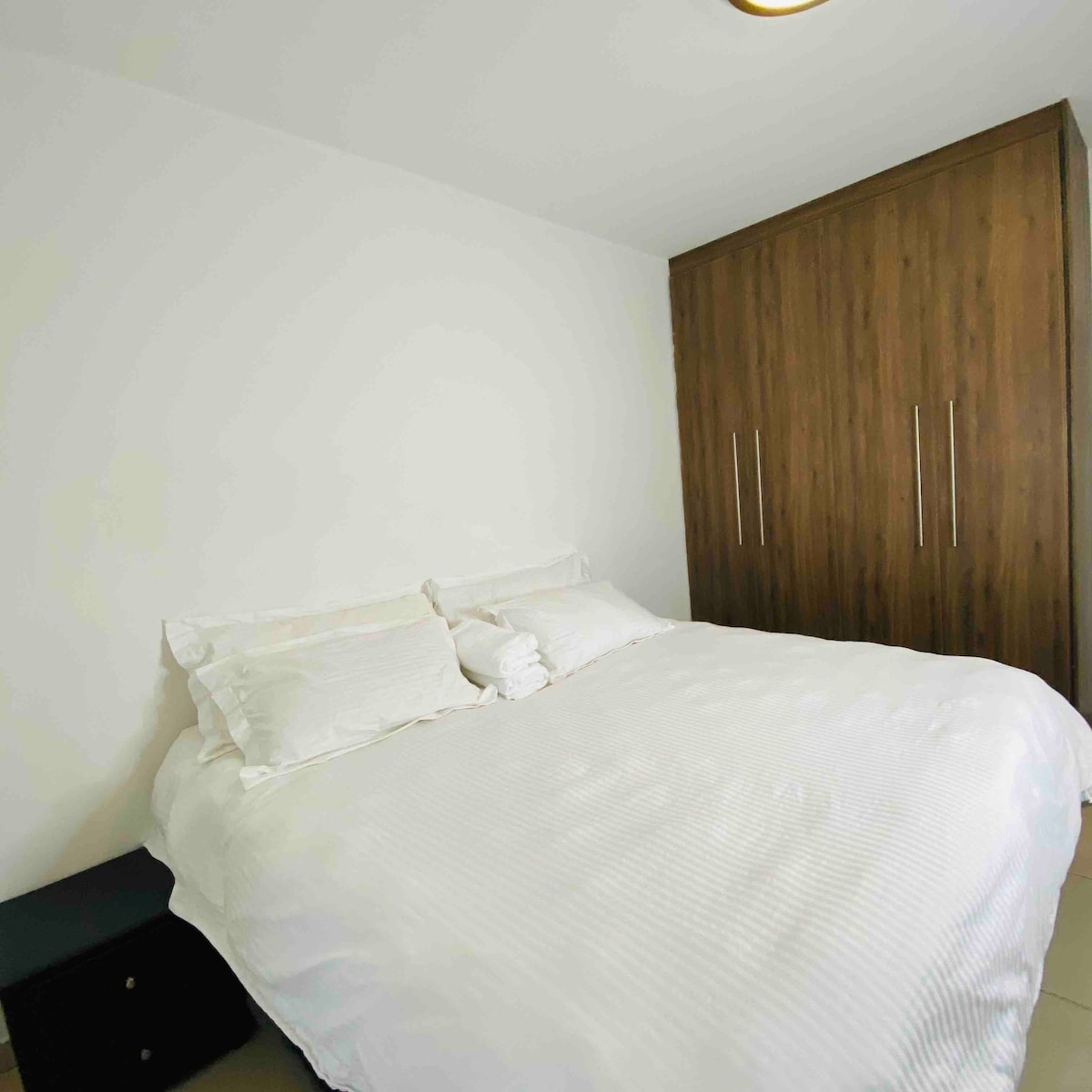 A well-lit bedroom features a spacious bed with white linens and a pair of pillows. A dark nightstand is positioned beside the bed, while a tall wooden wardrobe with multiple doors stands against the wall, contributing to a clean and minimalistic design.