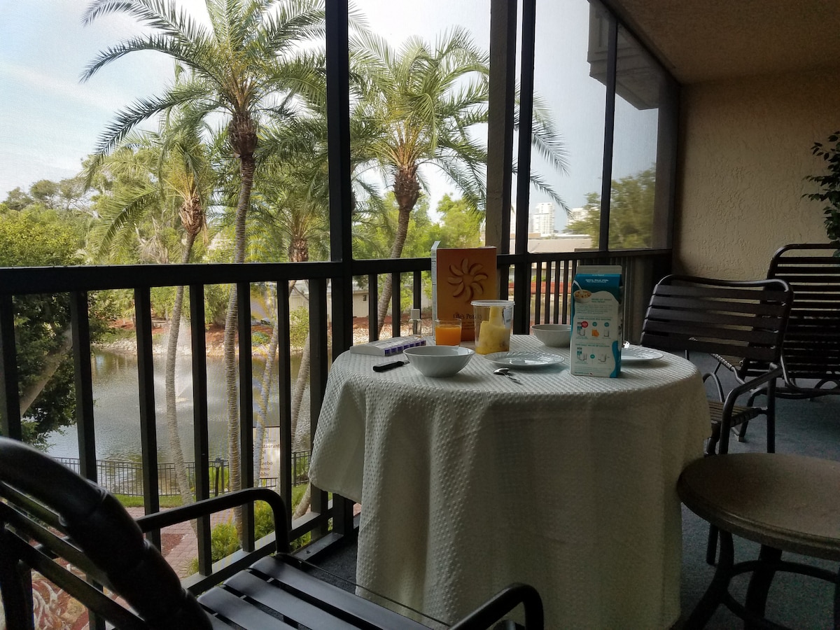 A lanai table is set with breakfast items, including bowls and a juice carton, facing a tranquil pond view with palm trees in the background. Natural light filters through the screened enclosure, enhancing the serene atmosphere of the outdoor space.
