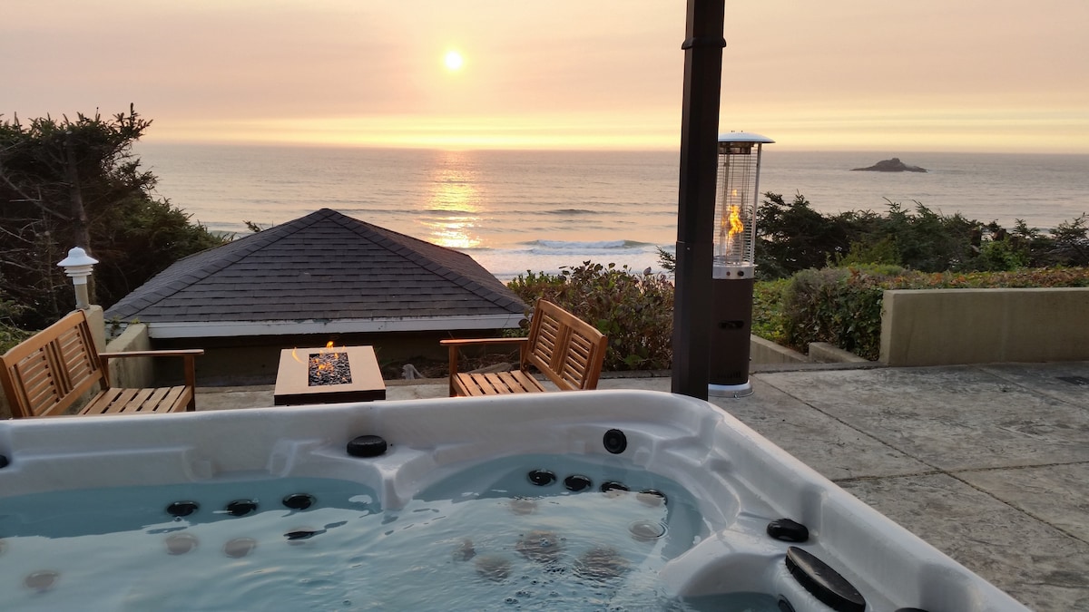 A hot tub sits on a stone patio, overlooking the ocean at sunset. A small fire table is positioned nearby, surrounded by wooden chairs. The view features gentle waves and an outcropping in the distance, while soft hues of orange and pink fill the sky.