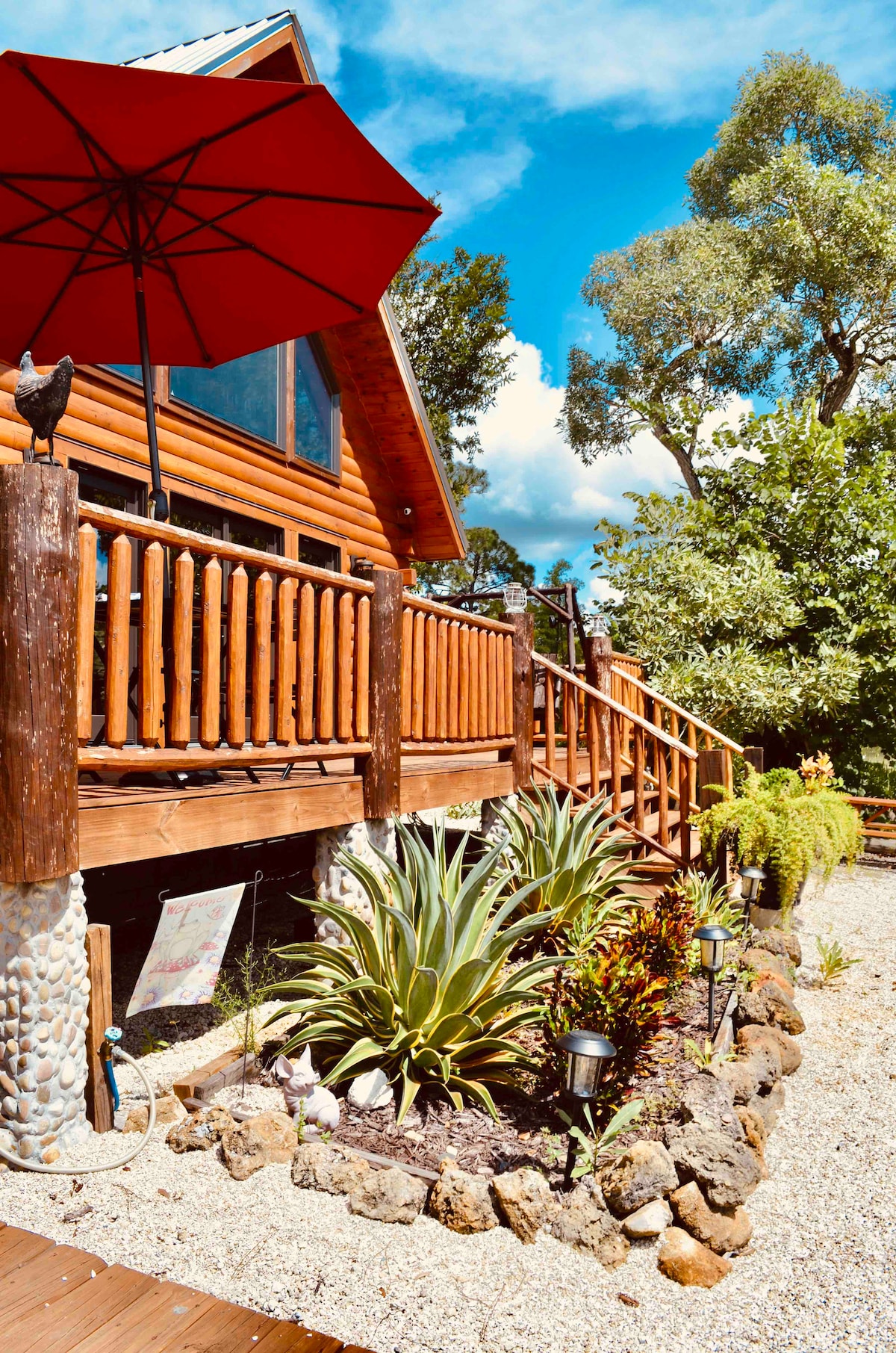 A charming log cabin features a wooden deck adorned with a vibrant red umbrella, providing shade. Lush greenery surrounds the property, with a landscaped garden showcasing various plants and stones, creating a welcoming outdoor space. Large windows invite natural light into the home.