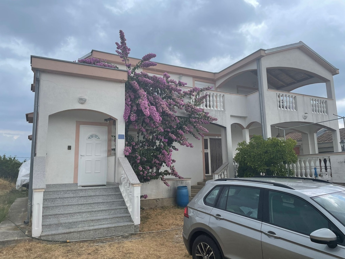 The exterior of a house is presented, featuring a staircase leading to the main entrance. Bougainvillea blooms in vibrant purple adorn the front, contrasting with the light-colored walls. A parked car is visible in the foreground, while a covered balcony is situated on the upper level.