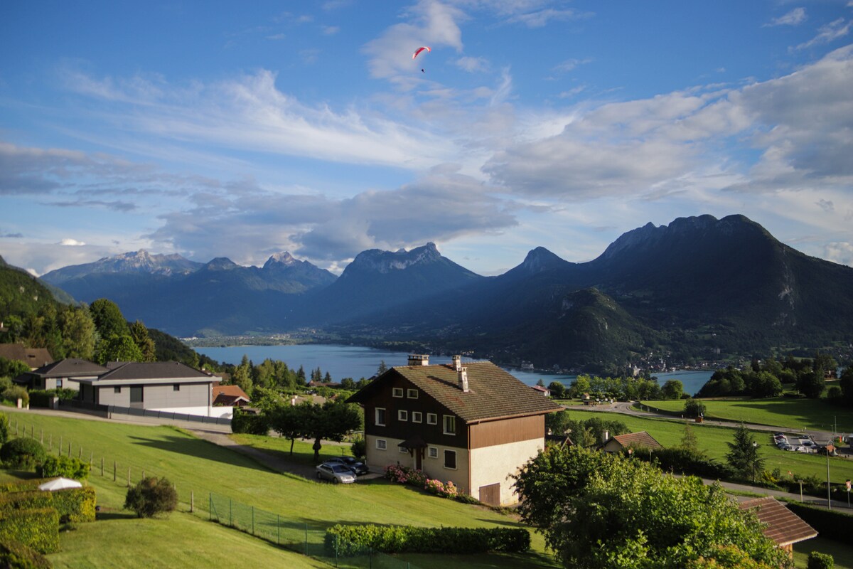 A serene landscape is captured, featuring rolling green hills and a view of a lake bordered by distant mountains. A traditional house is positioned in the foreground, with a yard lush in greenery. A paraglider is seen soaring gracefully in the clear blue sky.