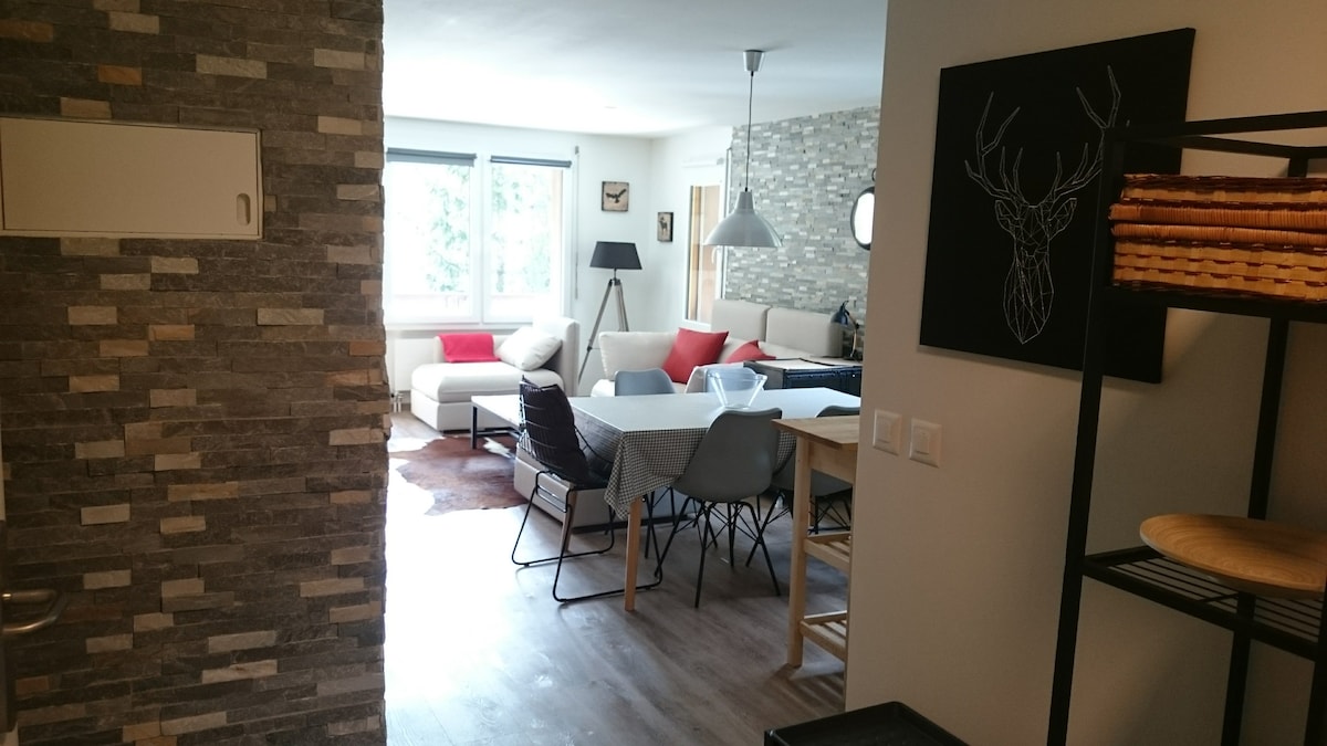 A modern open-plan living area is visible, featuring a spacious seating arrangement with a large white sofa and colorful throw pillows. A dining table with chairs accompanies light wooden elements, while stone accents provide a rustic touch to the overall design.