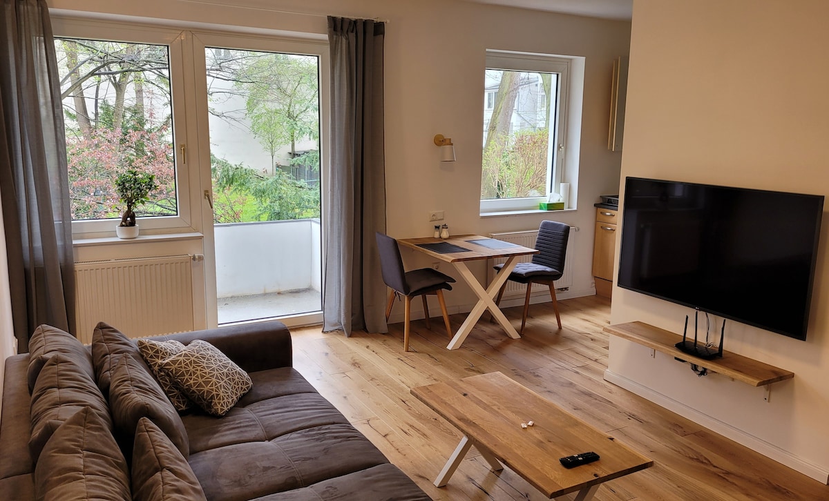 A spacious living area is showcased, featuring a large sofa, a wooden coffee table, and a flat-screen TV mounted on the wall. A dining table for two is positioned near the windows, which frame a view of the greenery outside.