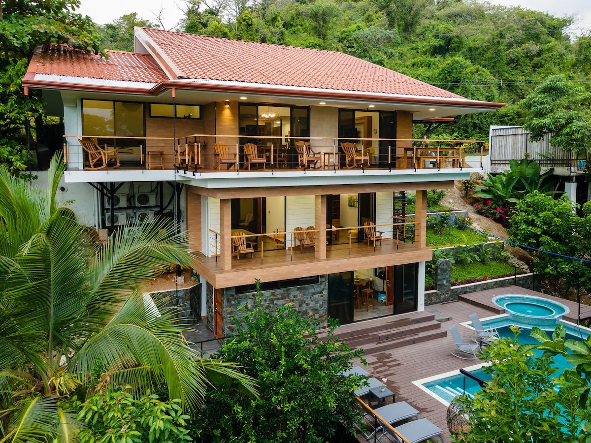 A modern two-story villa showcases a spacious wooden deck and multiple balconies surrounded by lush greenery. Below, a private pool is framed by lounge chairs. The architecture features large windows, blending with the natural landscape.