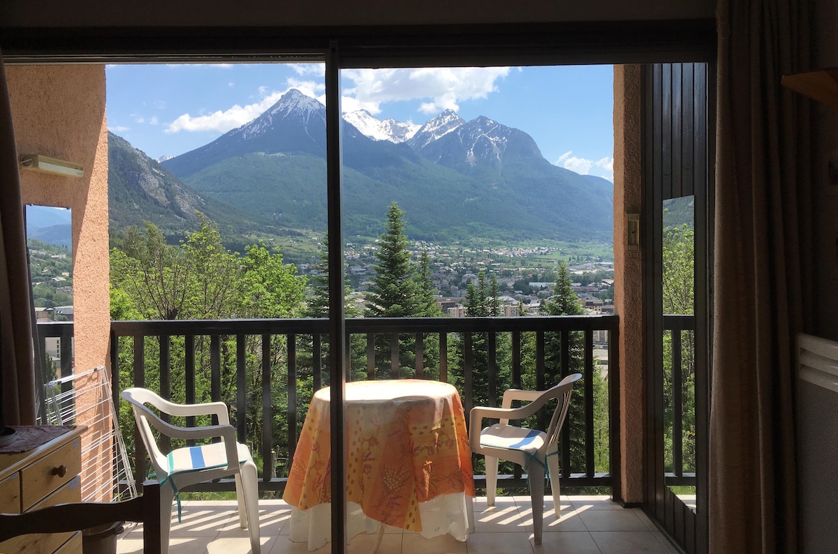 A balcony view showcases lush greenery and mountains beyond, framed by large glass doors. Two white chairs and a round table with a floral tablecloth are positioned on the tiled floor, inviting relaxation while enjoying the scenic landscape.