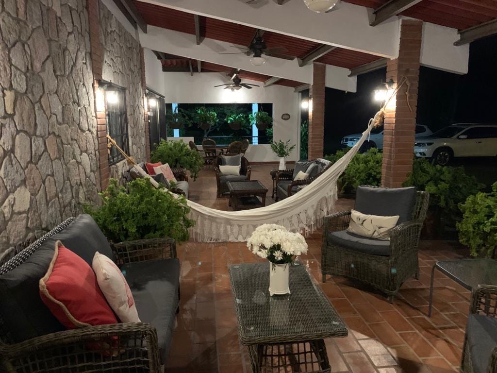 A spacious covered porch features a hammock alongside comfortable seating arrangements, including wicker chairs and a glass-top table. Soft lighting illuminates the area, and greenery surrounds the seating, creating a relaxing atmosphere. The view beyond showcases a tranquil exterior area.
