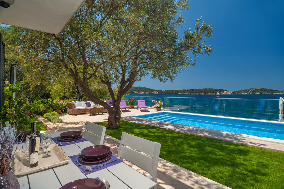An outdoor dining area features a long white table set with dark plates and glasses. Lush green grass surrounds the table, complemented by a shady olive tree. A private pool glimmers under the sun, with sun loungers positioned nearby, offering views of the tranquil sea.