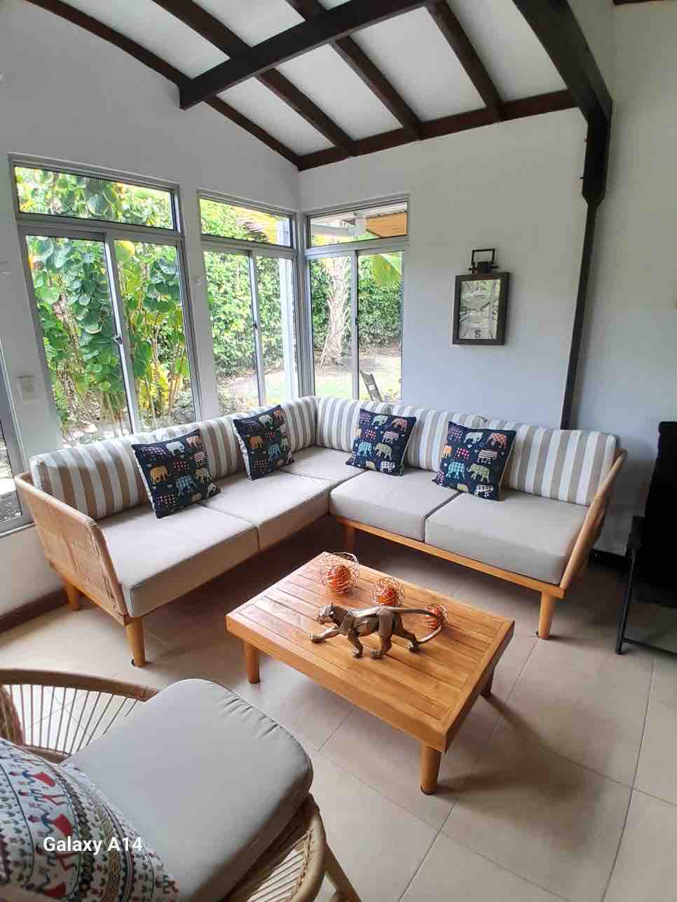 A cozy sitting area is equipped with a curved sofa adorned with elephant print cushions. A wooden coffee table sits in the center, complemented by decorative items. Large windows allow natural light to fill the space, providing a view of the surrounding greenery.