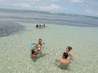 A shallow coastal area is shown, with several people enjoying the clear water. Gentle waves can be seen in the distance, creating a serene atmosphere. The sandy bottom is visible, complementing the light blue and green tones of the water under the bright sky.