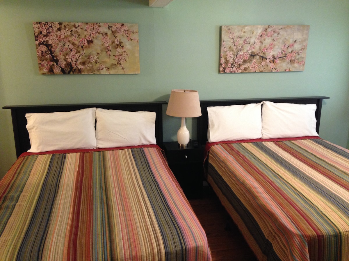 The bedroom features two spacious beds, each adorned with colorful striped bedding. A nightstand with a lamp is positioned between the beds. Cherry blossom artwork hangs above the beds, complementing the soft green walls.
