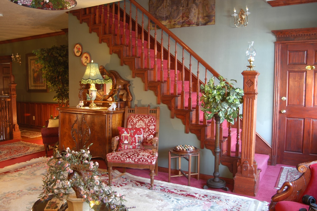 An elegant entryway features a grand staircase with richly detailed wooden railings. A vintage lamp illuminates a wooden side table, accompanied by a decorative armchair and potted plants. The space is adorned with area rugs, creating a warm and inviting atmosphere.