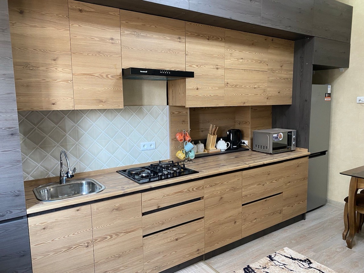 A modern kitchen features wood cabinetry, complemented by a light-colored countertop. A gas stove with an overhead exhaust hood is positioned alongside a microwave. A small sink and essential appliances are neatly arranged, creating a functional cooking space. Decorative items add a subtle touch.