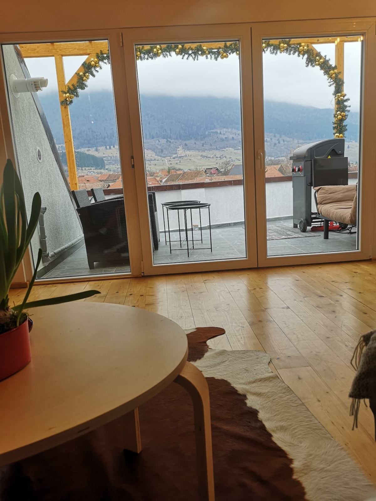 A spacious room features a large glass door leading to a private terrace adorned with greenery. Outside, mountains can be seen in the distance. Inside, a round table and a cozy seating area are complemented by a decorative rug on the wooden floor.