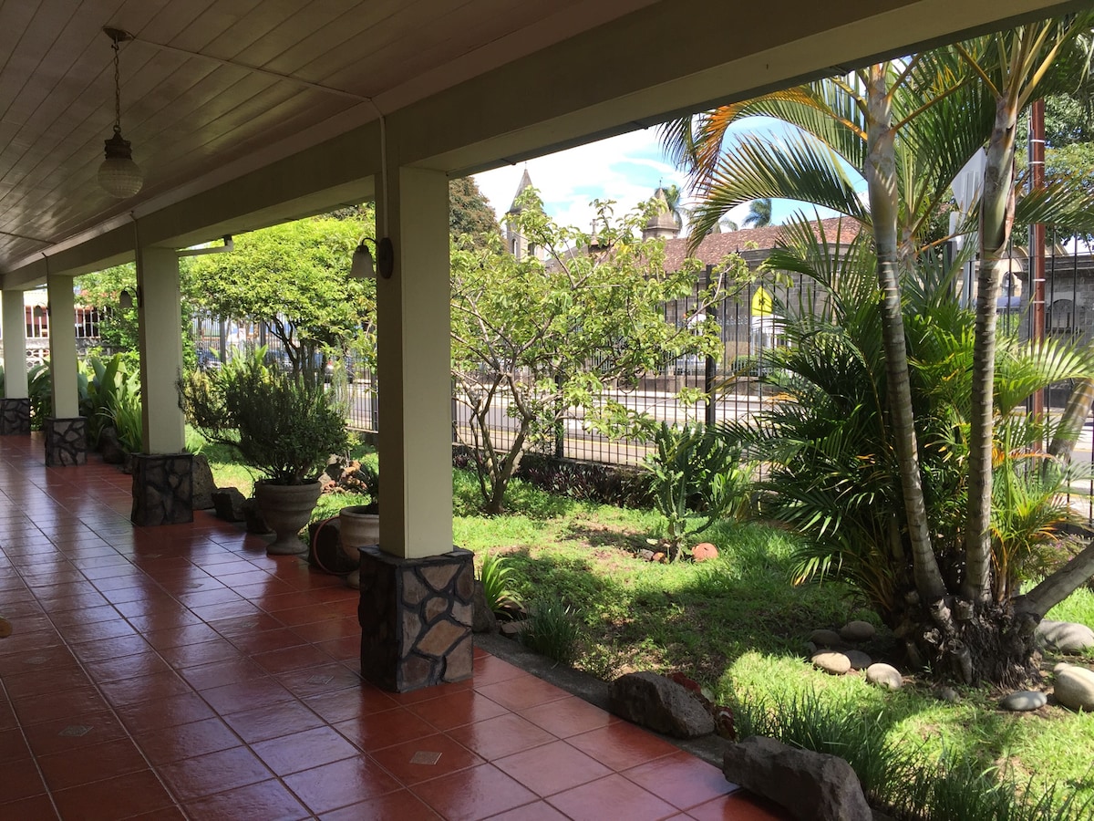 A covered terrace is presented, featuring terracotta tile flooring. Lush greenery is visible beyond decorative stone planters, with sunlight filtering through palm leaves. The view includes a peaceful street scene, enhancing the tranquil outdoor atmosphere.