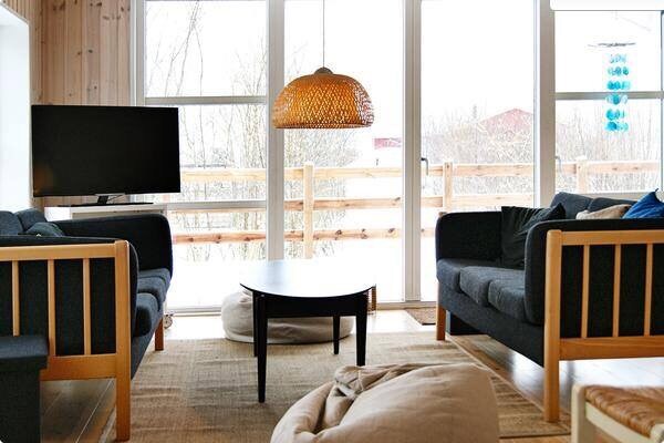 A cozy living area features two dark sofas and a low coffee table, creating an inviting space for relaxation. Natural light floods in through large windows, accented by a warm pendant light overhead. The soft texture of floor cushions complements the wooden flooring.