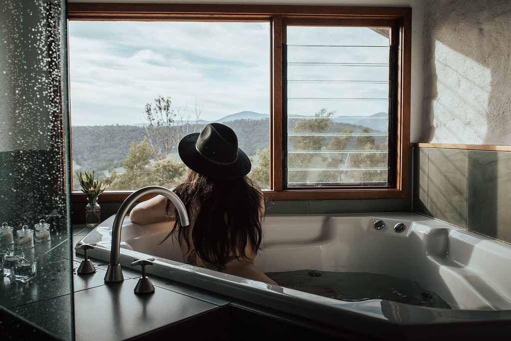 A spacious double spa bath is positioned beside a large window, offering expansive views of the surrounding mountains and nature. Natural light fills the room, and a small plant rests on the windowsill, enhancing the serene ambiance of the private space.