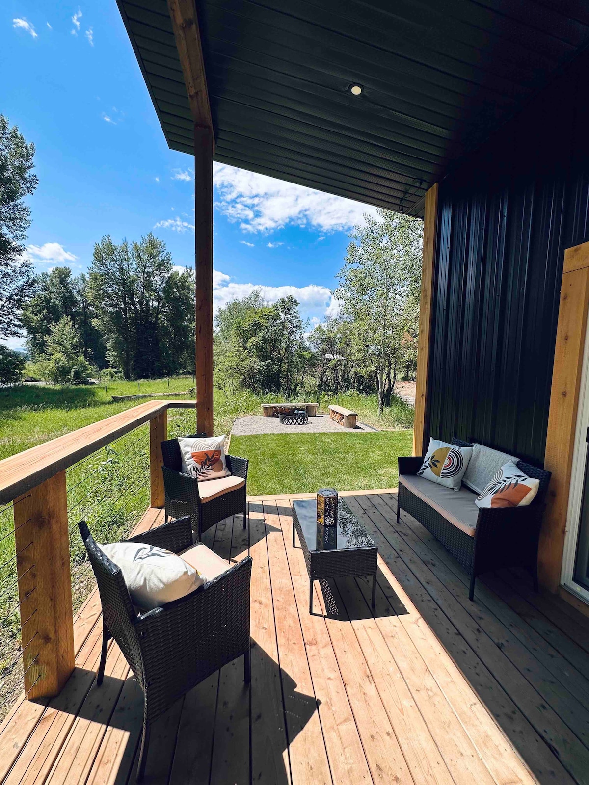 A private deck area features two wicker chairs and a small table, overlooking a green yard with trees. A fire pit is visible in the distance, surrounded by seating. Natural light fills the space, enhancing the connection to the outdoors.