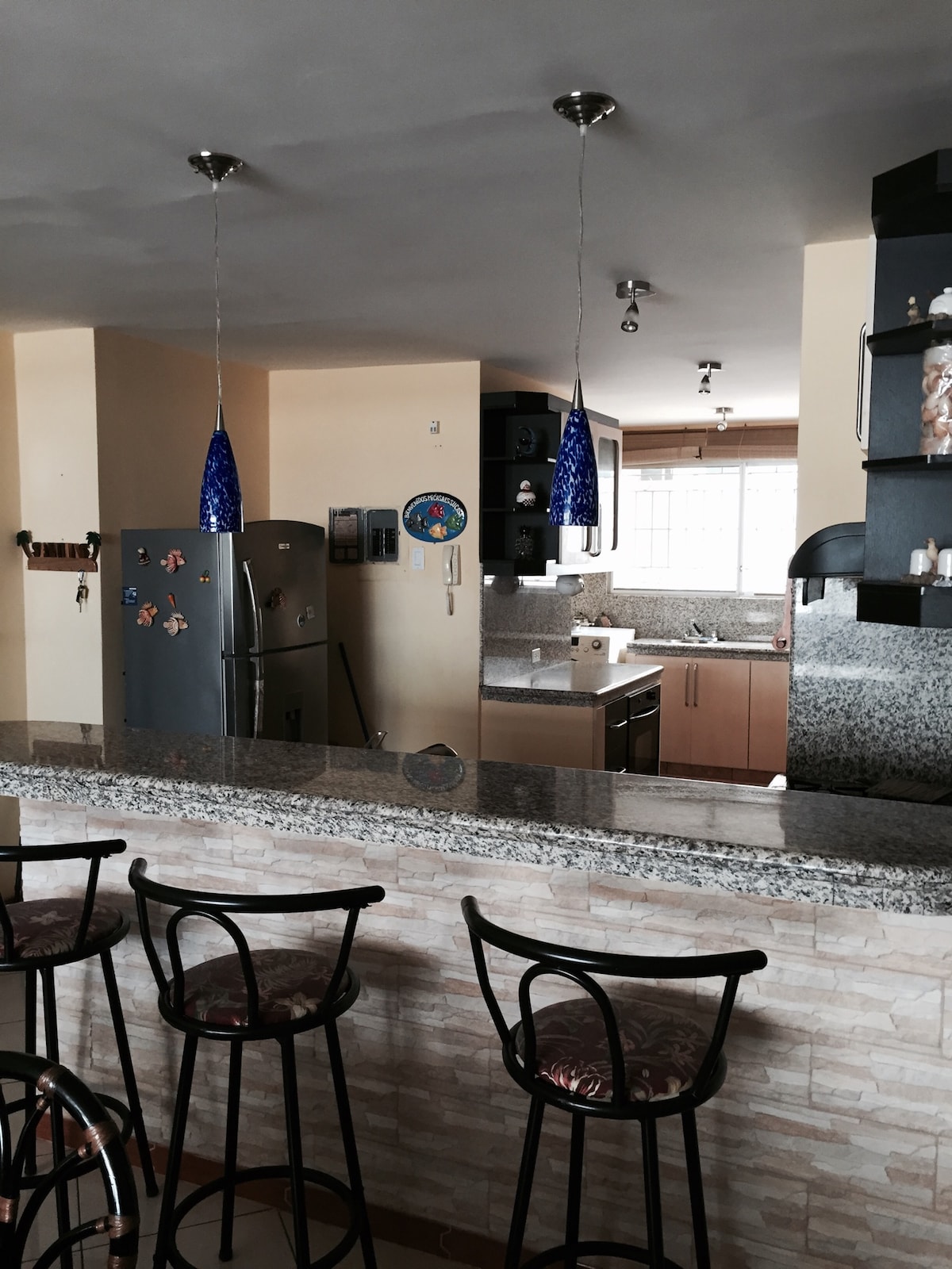 A modern kitchen area showcases a spacious countertop with bar seating for four. Bright blue pendant lights hang above, complementing the stainless steel appliances. A window allows natural light to illuminate the room, highlighting the sleek cabinetry and granite surfaces.