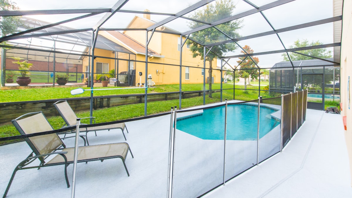 A screened pool area features a turquoise swimming pool surrounded by a spacious deck. Two lounge chairs are positioned for relaxation, with greenery visible beyond the enclosure. The outdoor space is designed for privacy and comfort.