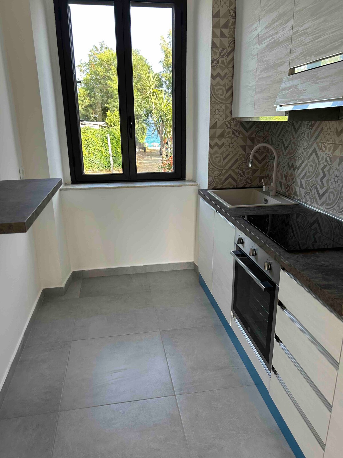 A modern kitchen features sleek cabinetry and a dark stone countertop. Natural light enters through a large window, offering a view of greenery outside. The kitchen is equipped with a built-in oven and a double sink, complemented by a patterned backsplash.
