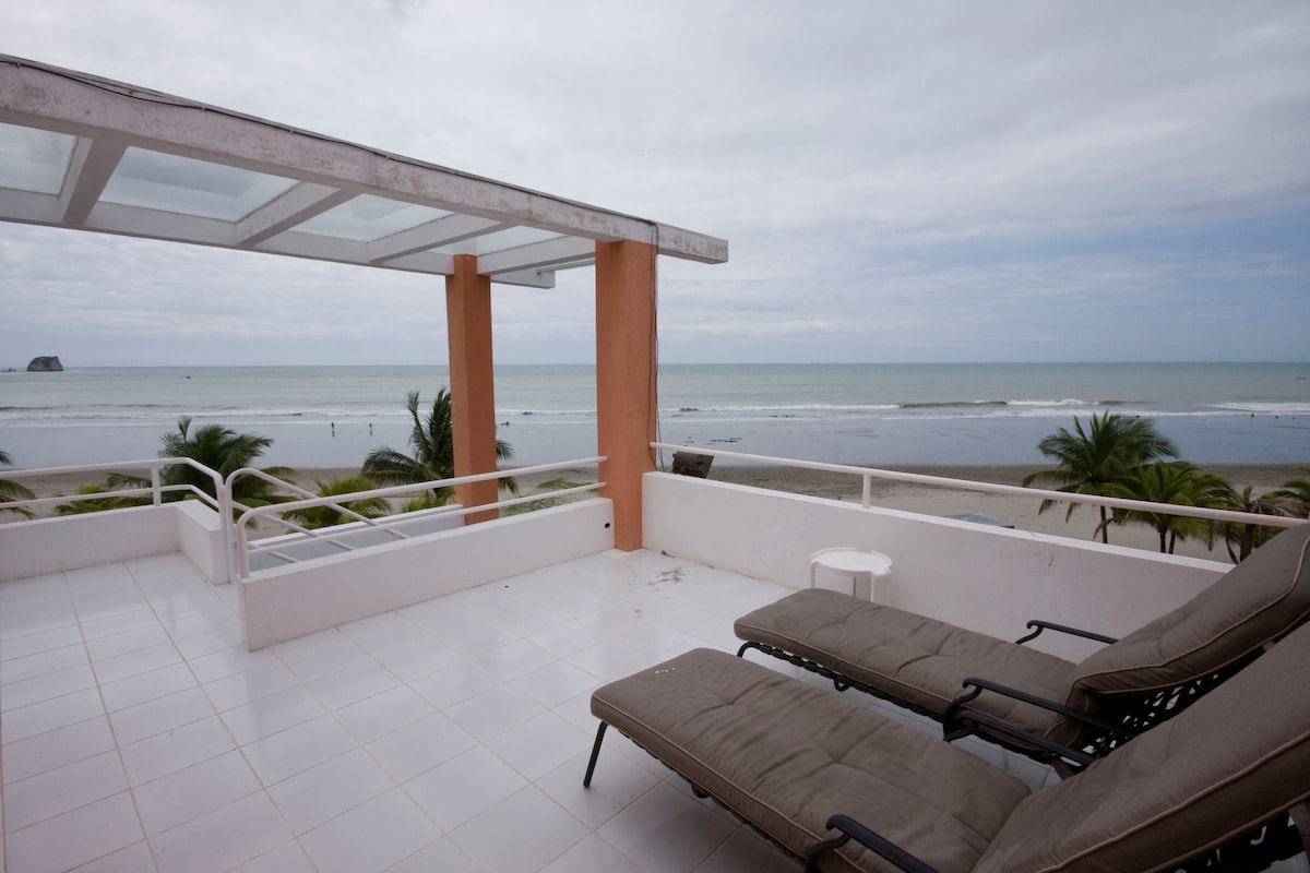A spacious terrace offers stunning ocean views, featuring two lounge chairs positioned for relaxation. The white tiled surface and surrounding palm trees enhance the open feel, while the horizon reveals gentle waves lapping against the shore.