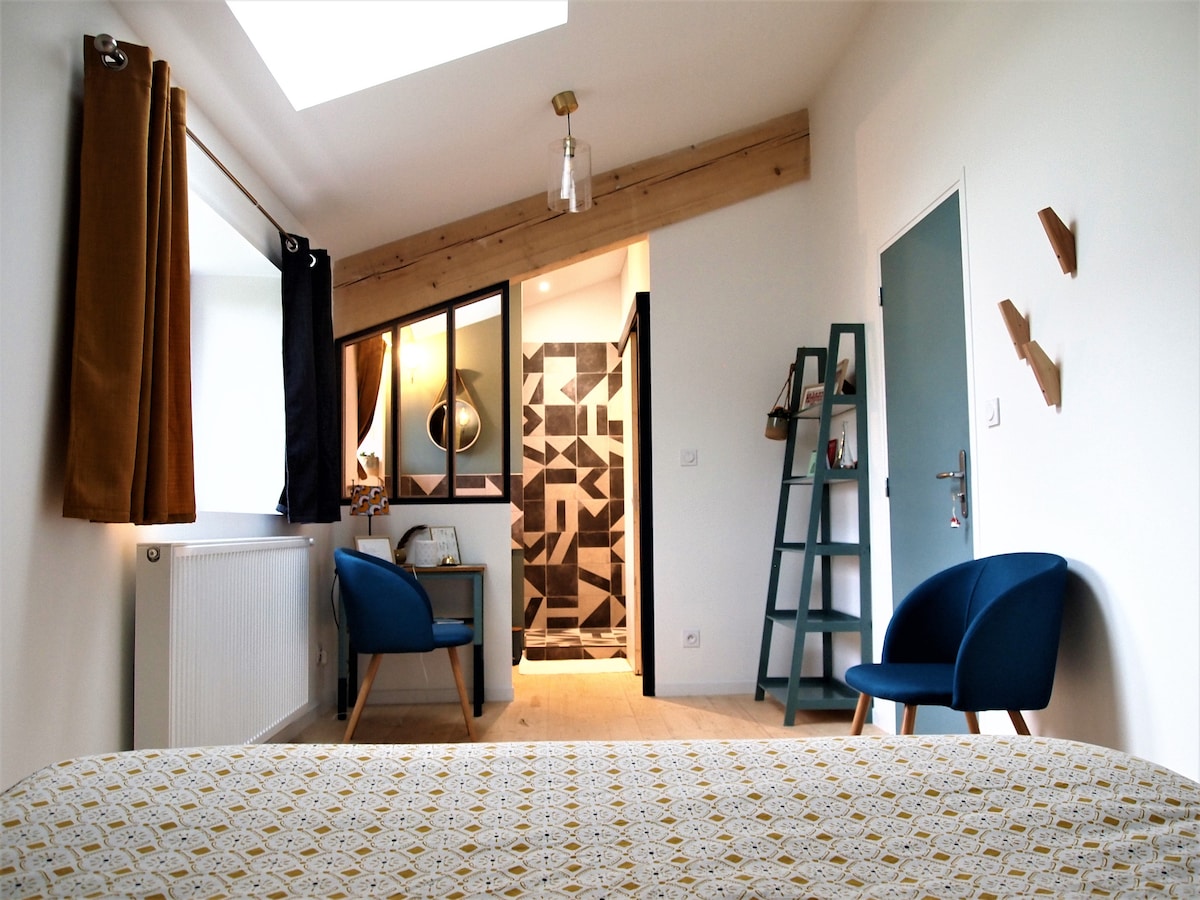The spacious room features a comfortable bed with a patterned coverlet. Two blue chairs are placed beside a small table. A well-lit bathroom with a modern design is visible through glass doors. Natural light enters through a skylight and a window draped with curtains.