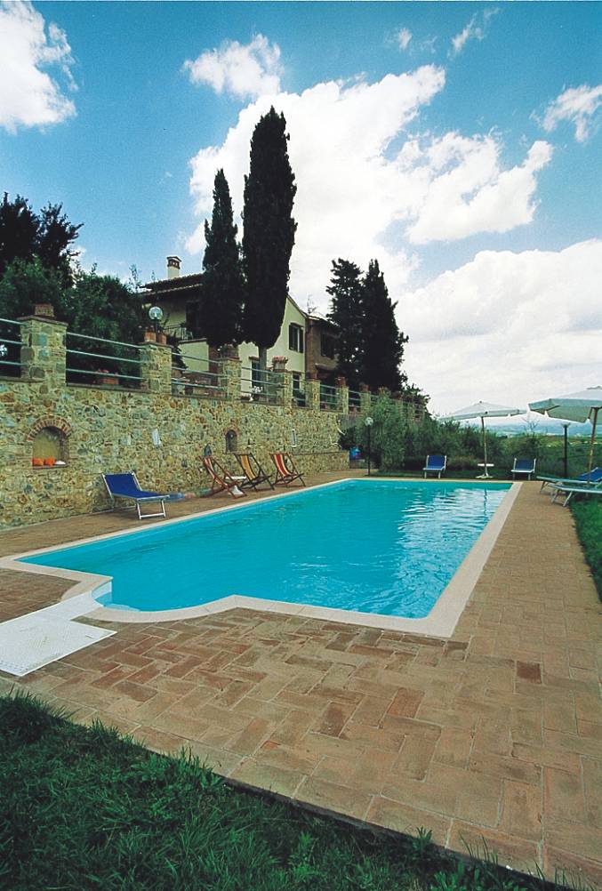 A shared swimming pool is set within a stone-walled area, surrounded by mature trees and olive groves. Several sun loungers are arranged along the poolside. The clear blue water reflects the sky, creating a tranquil outdoor space with a scenic view of the Tuscan landscape.