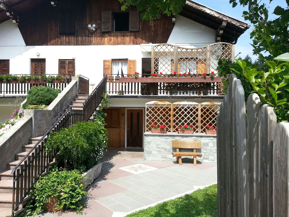 The exterior of the building features a charming alpine design, with wooden accents and flower boxes. A welcoming entrance is flanked by a well-maintained garden and a spacious terrace above, offering a pleasant outdoor space for relaxation.