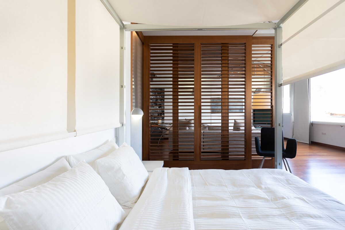 A modern bedroom features a spacious bed with soft white linens, positioned in front of large windows. A wooden partition with horizontal slats provides separation from the living area, which is visible in the background, enhancing the room's contemporary aesthetic.