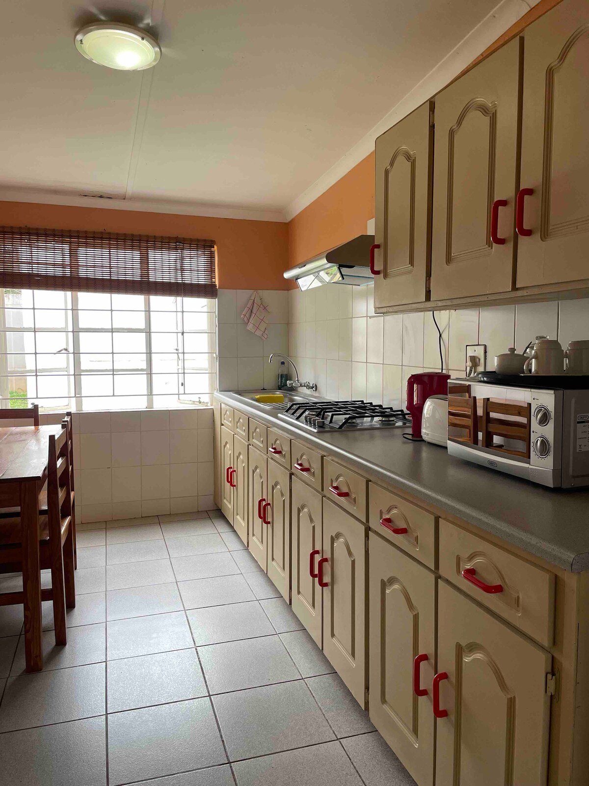 A functional kitchen space is featured with light beige cabinetry and gray countertops. A gas stove is positioned beneath a hood, with small appliances neatly arranged beside a window that provides natural light. A wooden dining table with chairs is visible in the corner.