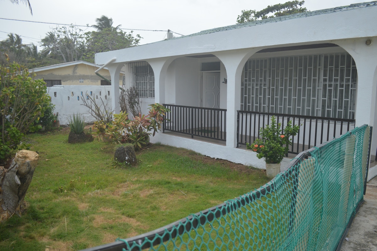 A residential exterior is shown, featuring a white-walled structure with arched entries. Green foliage and various plants are visible in the surrounding garden, complemented by a manicured lawn. A secure green fence outlines the property, enhancing the welcoming appearance.