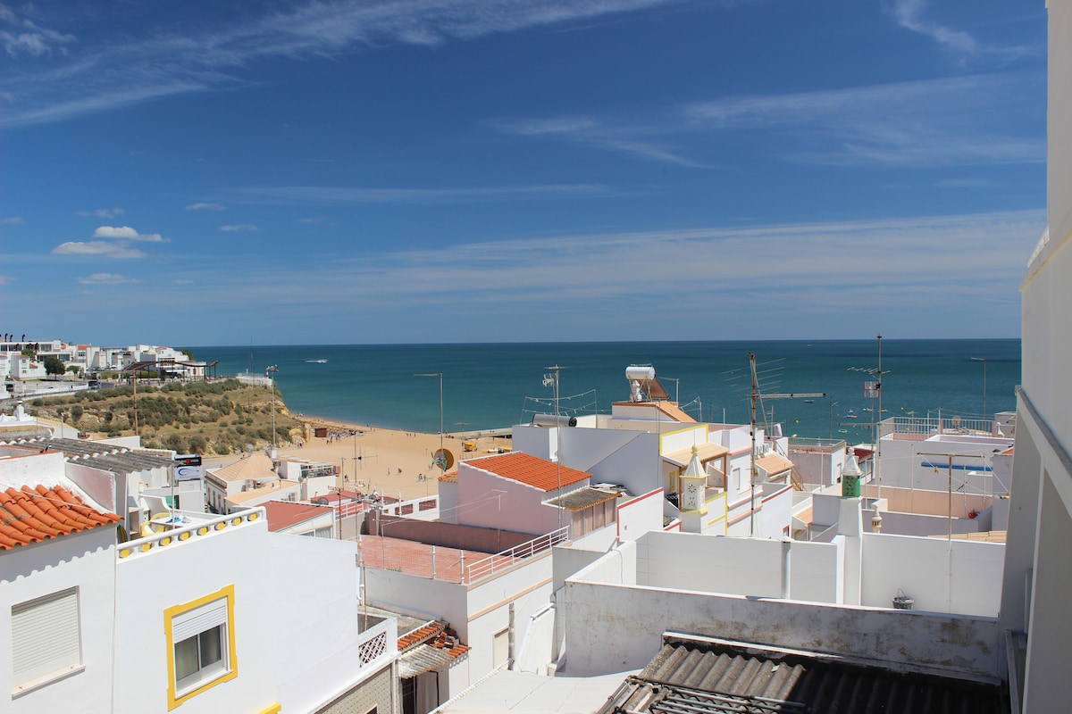 A panoramic view captures the vibrant rooftops of Albufeira, leading to the expansive beach below. The coastline stretches into the distance, framed by a clear blue sky with scattered clouds. The peaceful ambiance is complemented by the gentle waves lapping at the shore.
