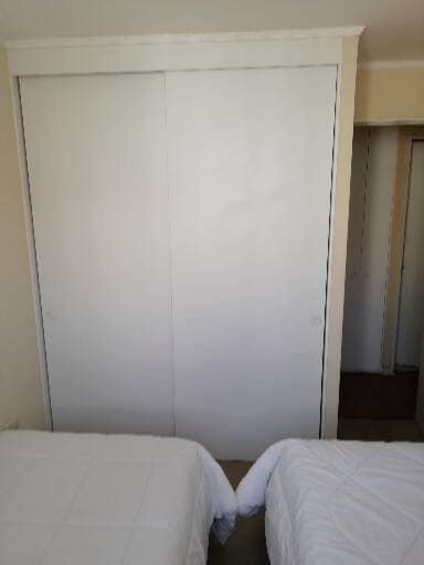 A minimalist bedroom features two neatly made beds with white bedding, positioned side-by-side. A large, double sliding closet door occupies the wall, enhancing the room's clean and spacious appearance. The subtle color palette contributes to a calm environment, with a glimpse of an adjoining room visible.