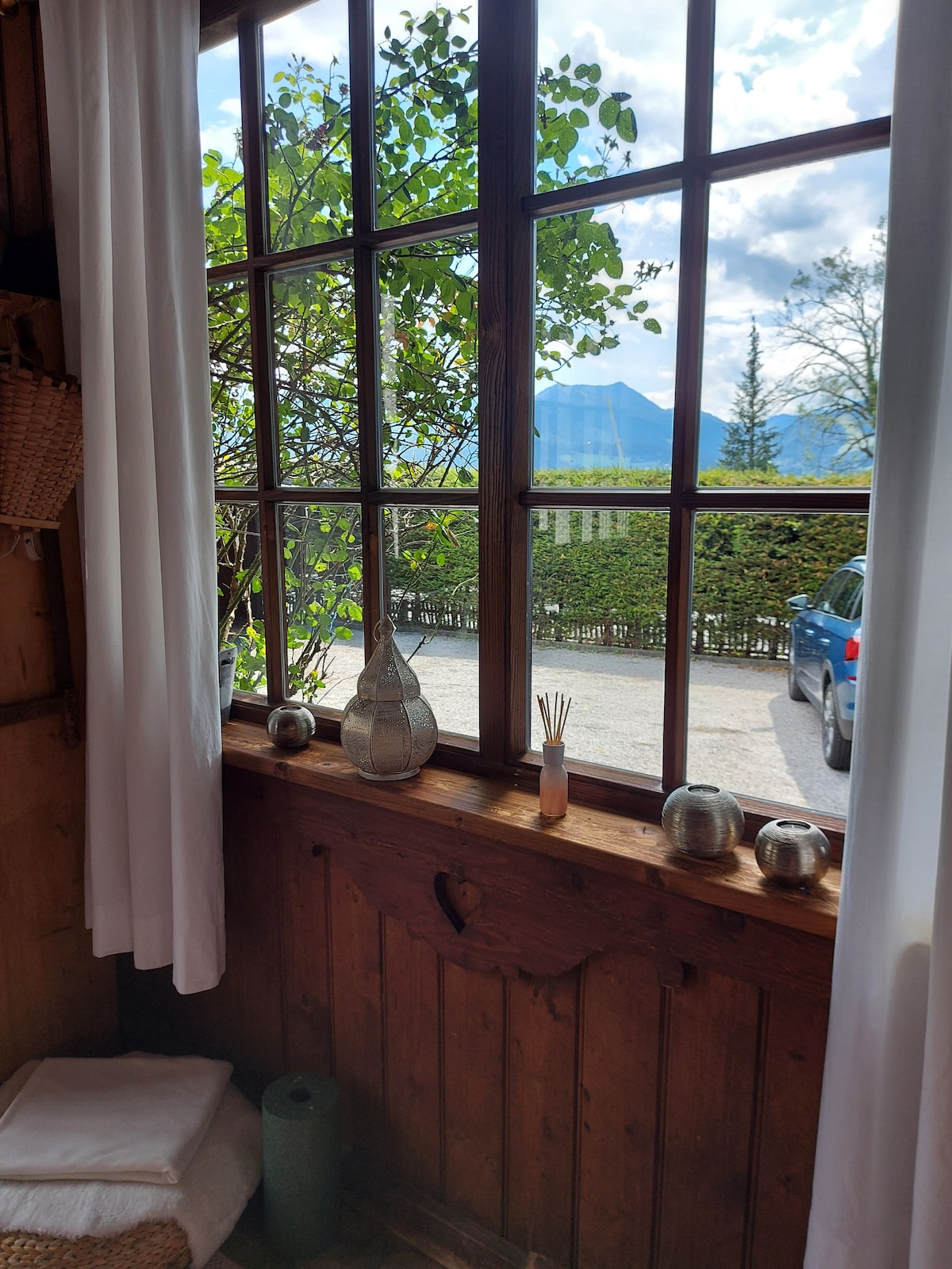 A wooden window frame provides a view of the outdoors, where trees and distant mountains are visible. Decorative items, including a glass vase and aromatic reed diffuser, are positioned on the windowsill, complemented by soft curtains allowing natural light to enter the space.