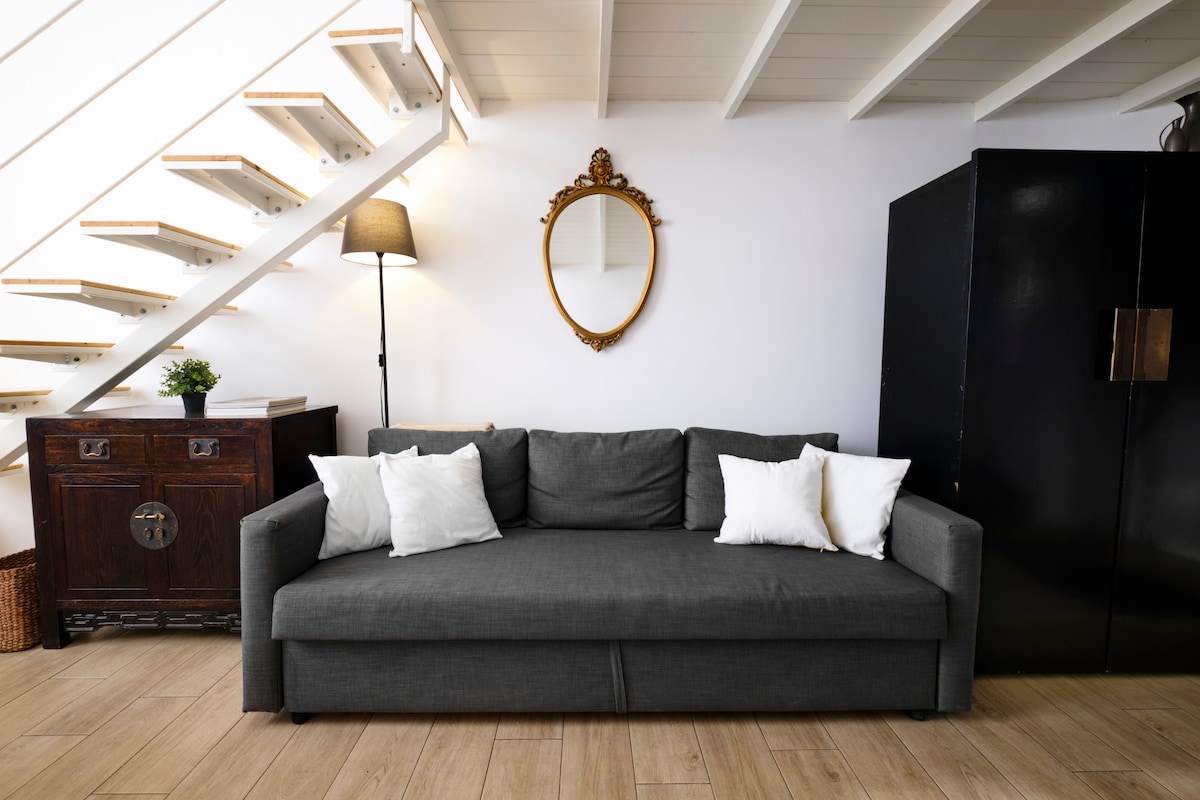 A cozy seating area features a dark grey sofa adorned with four white cushions. A wooden cabinet is positioned beside the sofa, and a standing lamp enhances the space. Above, a round mirror is framed in gold, reflecting the light from the nearby window.