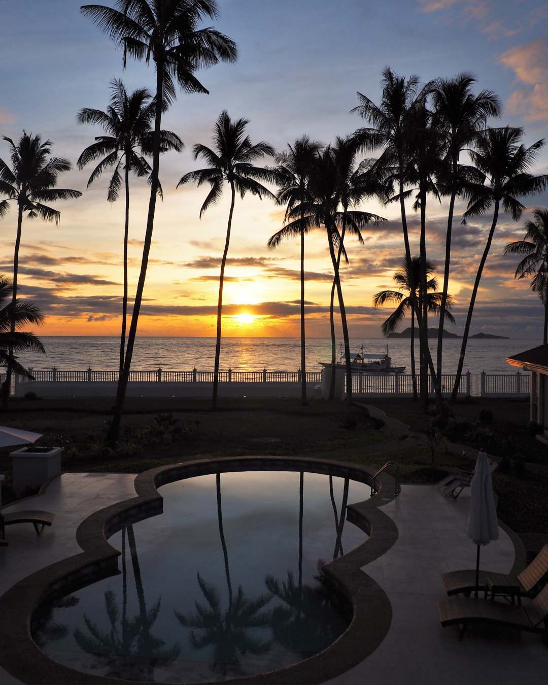A serene sunset is captured over a calm ocean, with palm trees framing the view. The reflection of the vibrant sky glimmers on the water surface of the chemical-free pool, creating a peaceful ambiance. Soft lighting enhances the scene as day transitions to night.