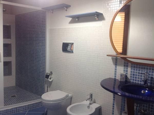 The private bathroom is equipped with a spacious shower, featuring glass doors and tiled walls in light blue hues. A toilet and bidet are present, while a round mirror above a blue sink adds a touch of color. Shelves provide additional storage above the sink.