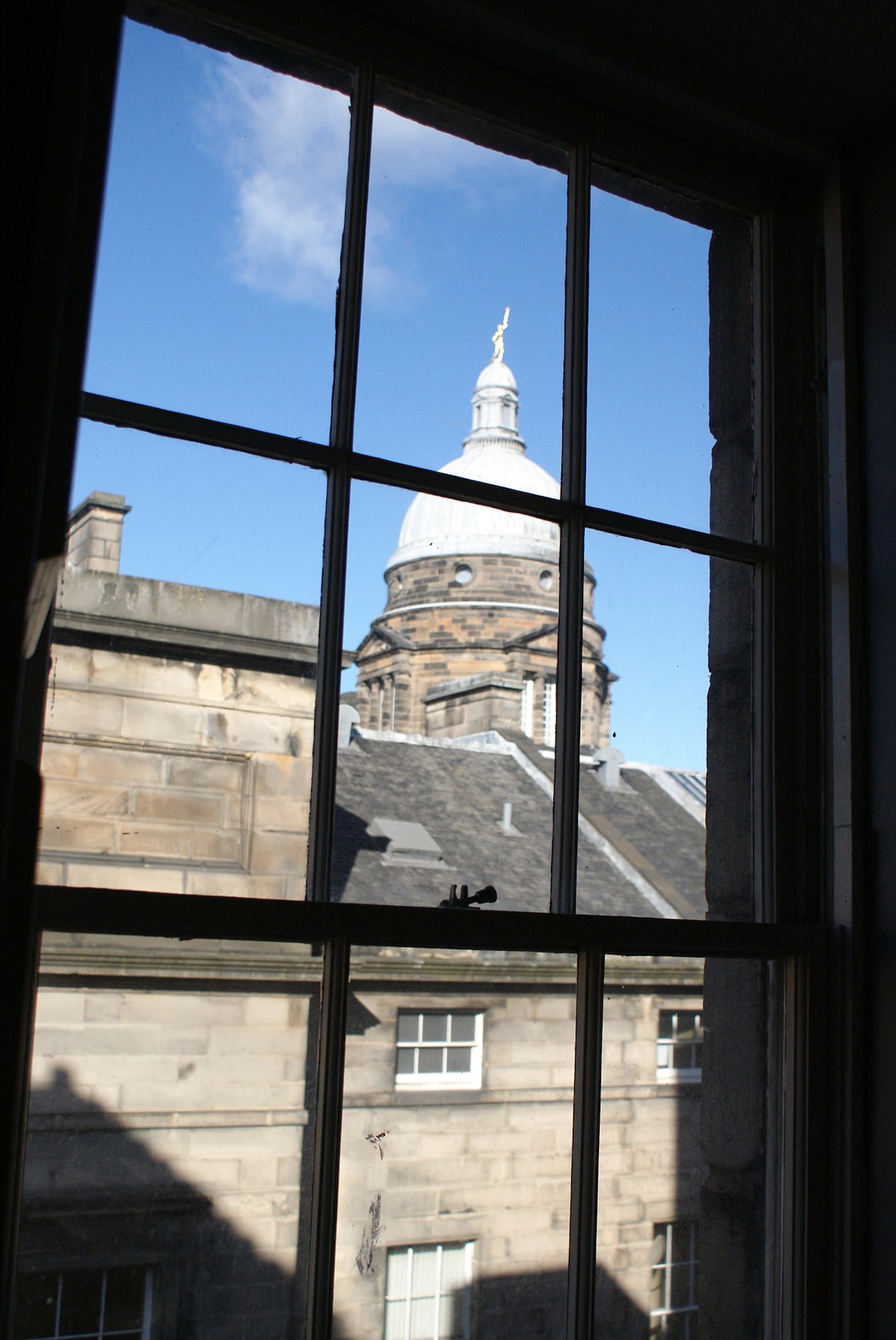 A view from the window reveals the dome of the Old College, showcasing its architectural detail against a clear blue sky. Stone buildings with slate roofs frame the scene, providing a glimpse of the historical charm of the surrounding area.