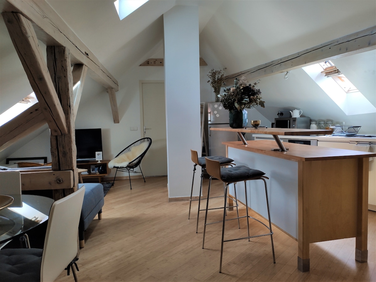 An open living space includes exposed wooden beams and a light-colored wooden floor. A small dining area is visible with stylish chairs. The kitchen, equipped with modern appliances, features a breakfast bar with stools. Natural light streams in through Velux windows on the ceiling.