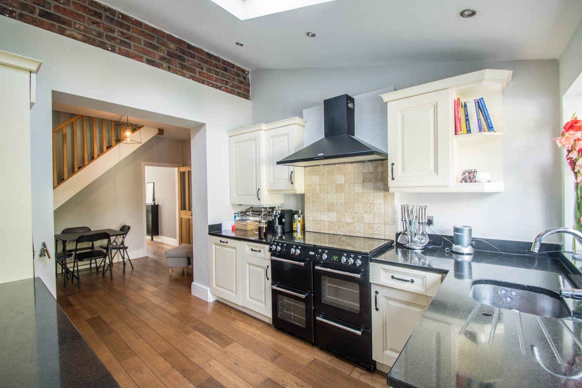 A modern kitchen with white cabinetry and sleek black countertops is showcased. A professional-grade stove and oven stand prominently, while open shelves hold cookbooks. Natural light floods in from a skylight, highlighting the wooden flooring and adjacent dining area visible in the background.