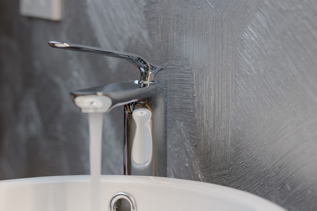A sleek chrome faucet is pictured, with a steady stream of water flowing into a white basin. The wall behind features a textured gray finish, adding a modern touch to the bathroom space.